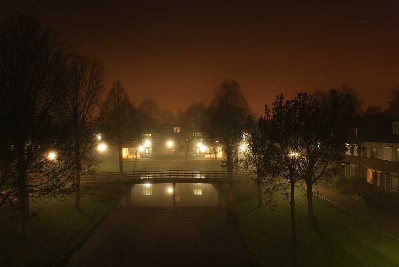 AA01.JPG - Een mooie mistopname hoeft niet altijd ergens in de "vrije" natuur te worden gemaakt. Ook in de bebouwing kan je mooie mistopnamen maken. Soms zelfs vanuit je huis. De belichting is net zo dat delampen van de straatverlichting net niet te fel zijn, maar dat er veel details zichtbaar zijn. De gelige lucht komt door de reflectie van natrium straatverlichting op de nevel.